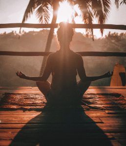 An out of focus picture of a girl with a top knot, sitting cross-legged, on a wooden deck, high up in the trees. You're seeing her from the back, the sun is just over her head and shining in your eyes, right through the leaves of a palm tree in front of her. I want to go to there!