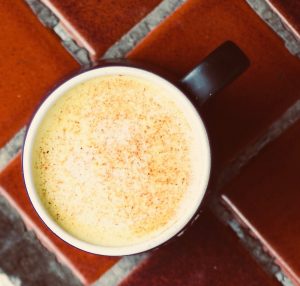 Brown ceramic mug full of turmeric orange colored golden milk, with spices floating atop, sitting atop pretty rust-colored kitchen counter tiles.