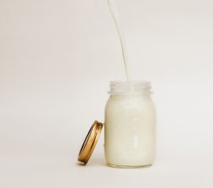 A stream of milk, flowing into a full mason jar, sitting on a white background, with a coppery-gold colored lid leaning up against it