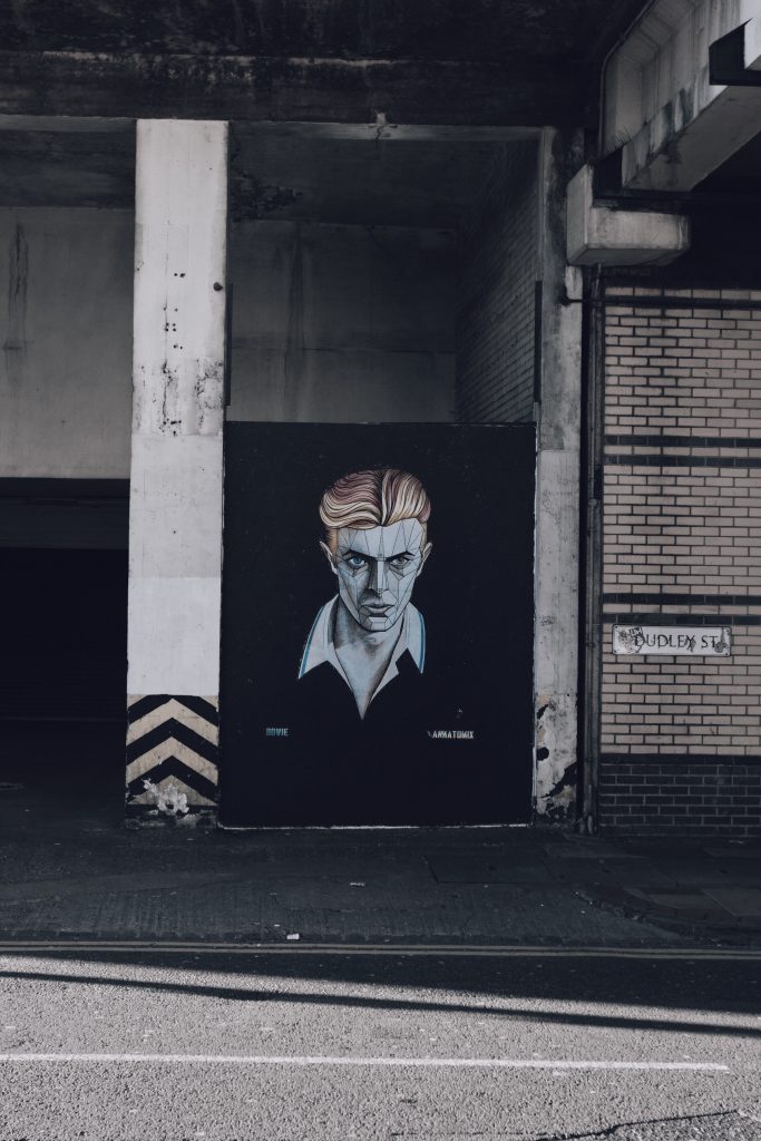 A  city street (the sign says "Dudley Street"), with a striking mural of David Bowie, Circa "Early 80's', looking dapper, menacing, and stylish.