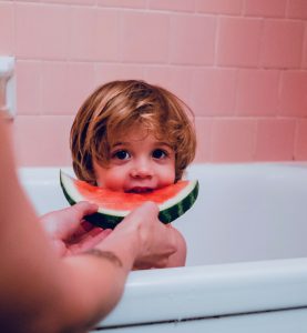 Tow-headed kid in a pink tiled bath being fed a piece of watermelon. Heaven!